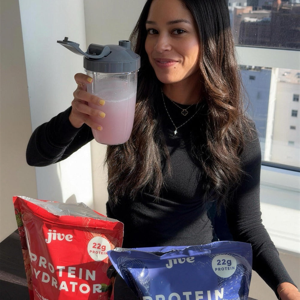 A woman holding a shaker of Protein Hydrator behind two bags